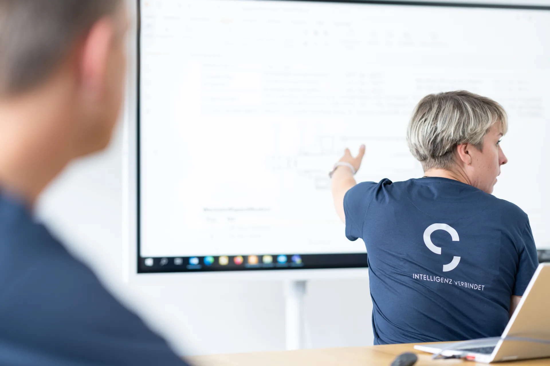 Eine Frau im dunkelblauen T-Shirt ist zu sehen, wie sie auf eine digitale Präsentationstafel zeigt und sich währenddessen redend zur rechten Seite dreht. Im Vordergrund ist verschwommen ein Schüler zu sehen.