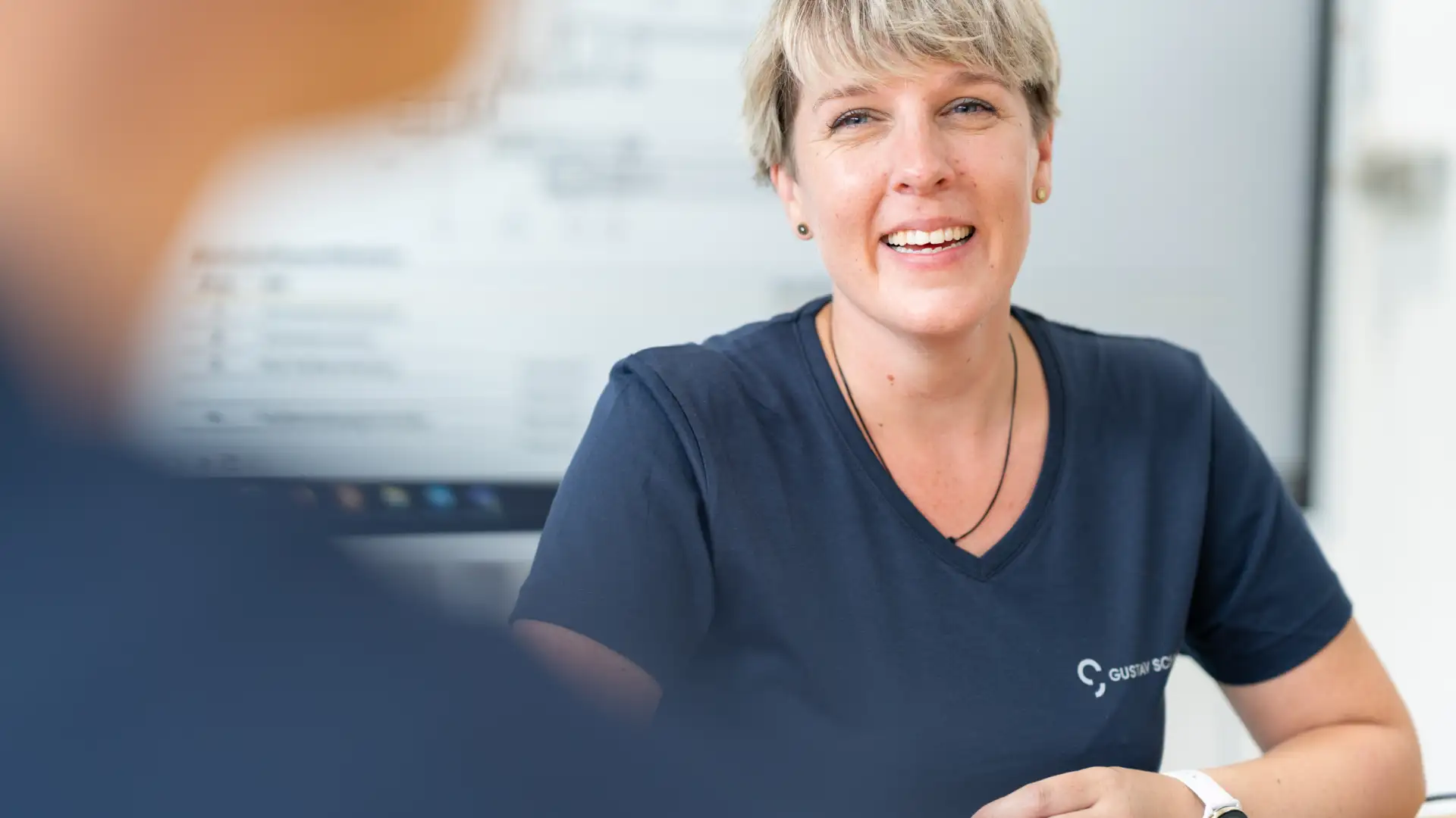 Frau mit blonden Haaren und blauem Gustav-Schmidt-Shirt schaut andere Person (nur verschwommen sichtbar) mit lachendem Gesicht an, im Hintergrund digitale Präsentationstafel zu sehen, am Tisch vor ihr ein Laptop und hat Stift in der Hand