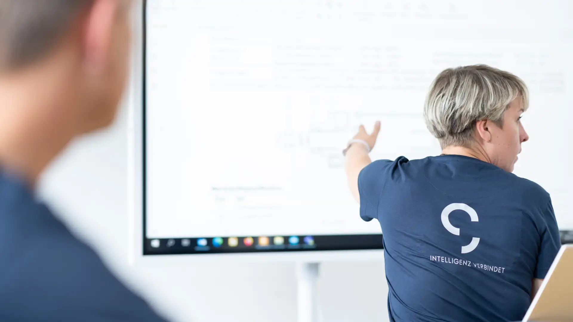 Eine Frau im dunkelblauen T-Shirt ist zu sehen, wie sie auf eine digitale Präsentationstafel zeigt und sich währenddessen redend zur rechten Seite dreht. Im Vordergrund ist verschwommen ein Schüler zu sehen.