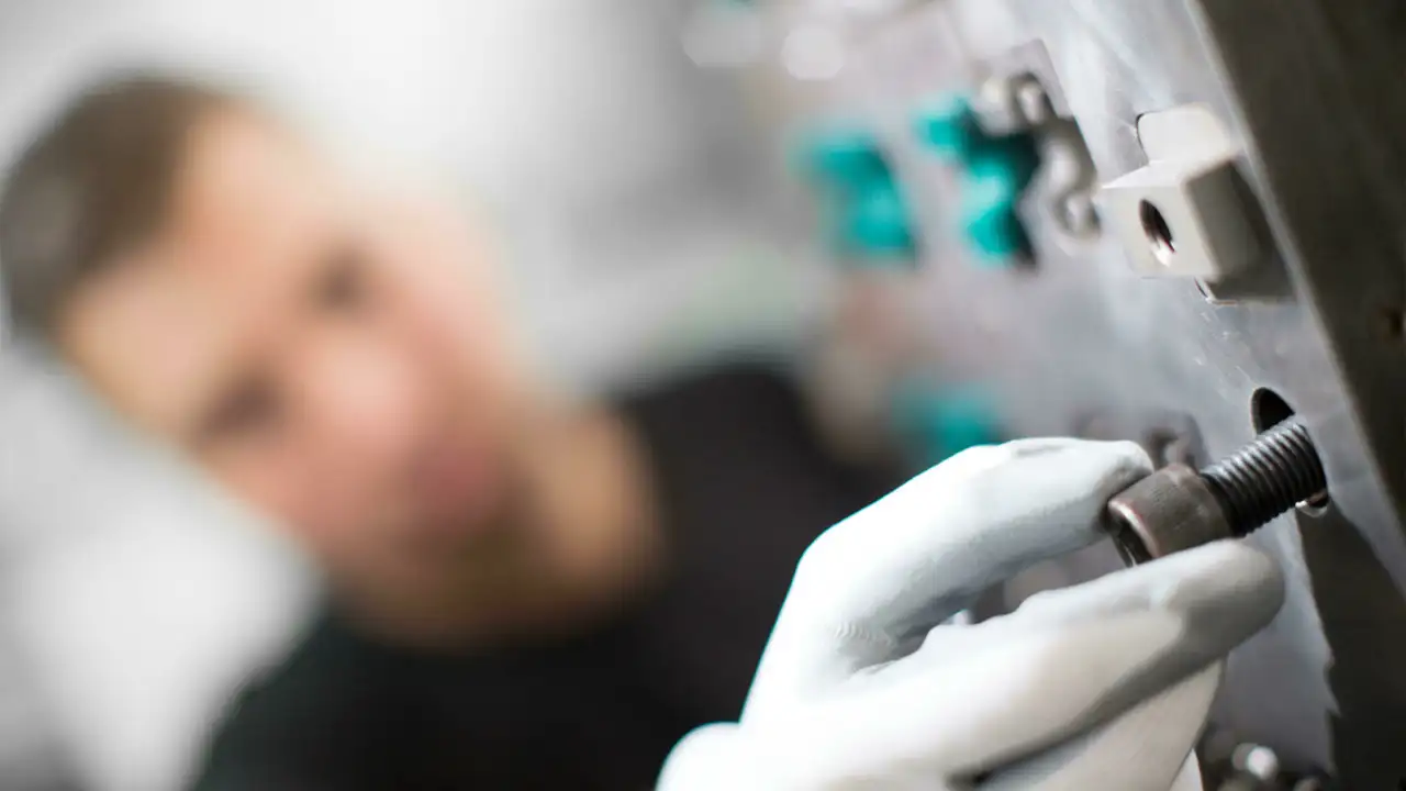 weißer Handschuh steckt Schraube in ein Loch in Maschine, im Hintergrund verschwommenes Gesicht von Mann erkennbar